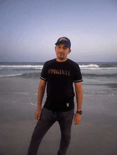 a man wearing a black shirt that says original stands on a beach
