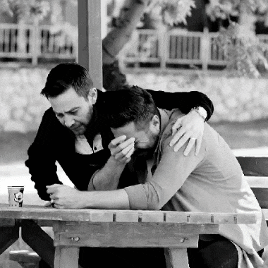 two men sitting at a picnic table with one covering his face