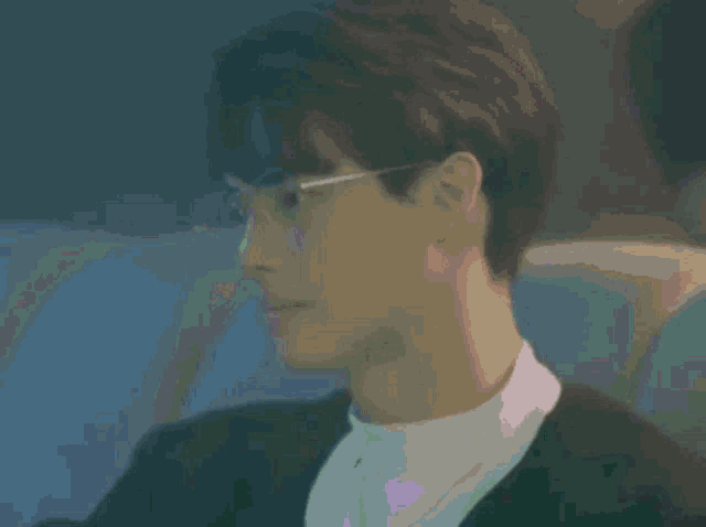 a young man wearing glasses and a black jacket is sitting in a dark room .