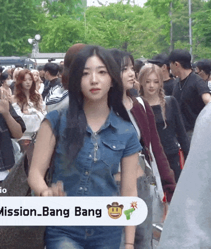 a woman in a denim shirt stands in a crowd with a mission bang sign behind her