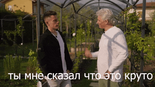 a man in a white shirt talks to another man in a black jacket in a garden