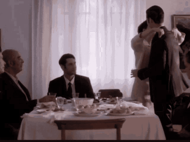 a man in a suit sits at a table with a woman in a white dress dancing in the background