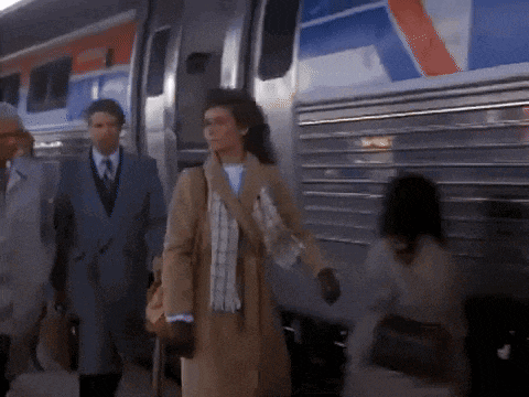 a woman in a trench coat is walking towards a train with a red white and blue stripe on the side