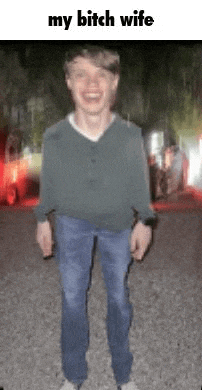 a young man in a green sweater and blue jeans is standing in a gravel driveway .