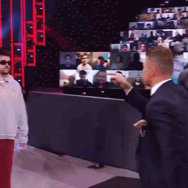 a man in a suit and sunglasses is standing in front of a screen with a group of people on it .