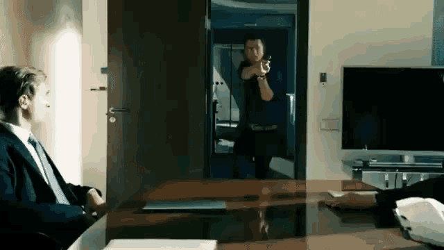 a man in a suit and tie is sitting at a desk while another man holds a gun