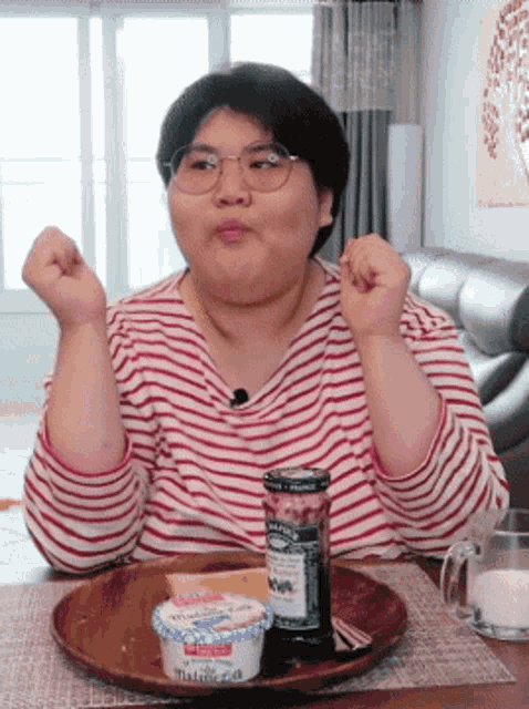 a woman wearing glasses is sitting at a table with a plate of food and a jar of jam