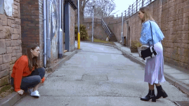 a woman squatting on the sidewalk next to a woman standing on the sidewalk