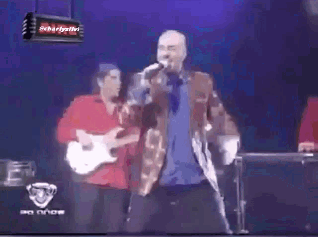 a man is singing into a microphone while another man plays a guitar behind him