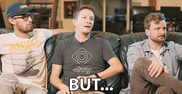 three men are sitting on a couch with one wearing a shirt that says oloy