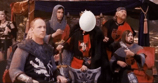 a group of medieval knights are sitting around a table with a man blowing a balloon in front of his face .