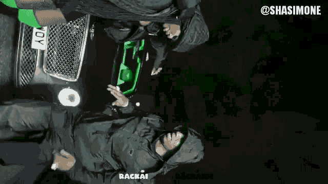 a man in a hooded jacket is standing in front of a green car with a license plate that says yoy
