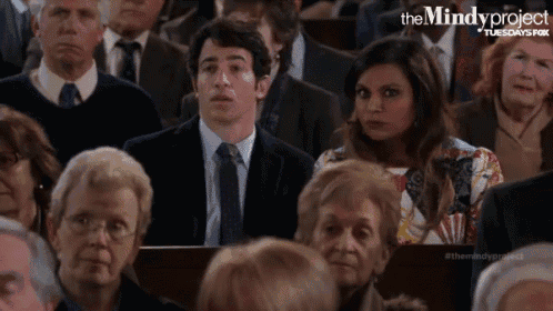 a man in a suit and tie is sitting in a crowd of people watching a presentation from the mindy project .