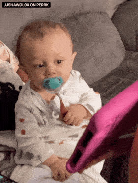 a baby with a pacifier is sitting on a couch with a pink tablet