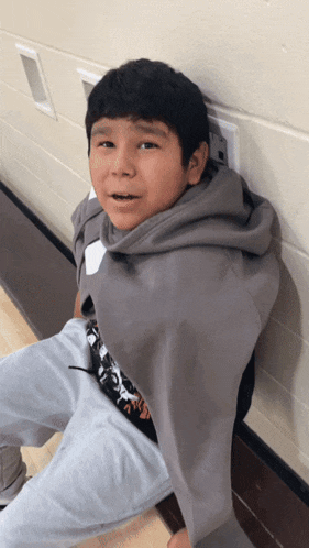 a young boy wrapped in a grey sweatshirt sits on a wooden floor