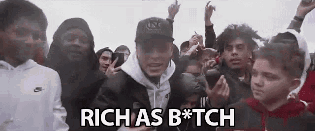 a man in a hat is standing in front of a crowd of people with the words rich as b * tch .