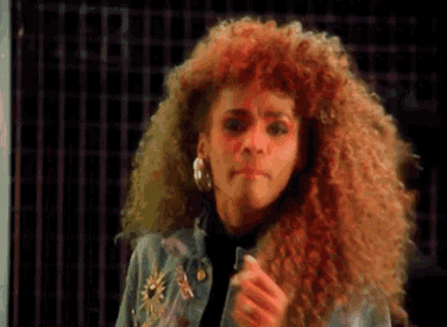 a woman with curly hair is wearing a denim jacket and earrings and making a funny face .