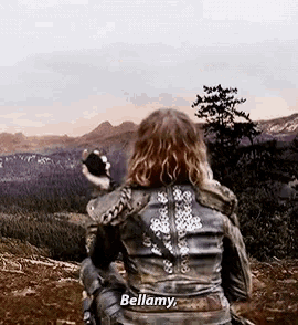 a woman in a leather jacket is standing on top of a hill holding a rock and says bellamy .