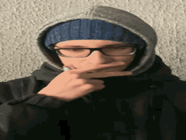 a man wearing glasses and a hooded jacket covering his face with his hand