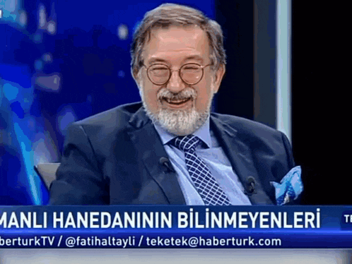 a man with glasses and a beard is sitting in front of a screen that says manli hanedanin bilinmeyenleri