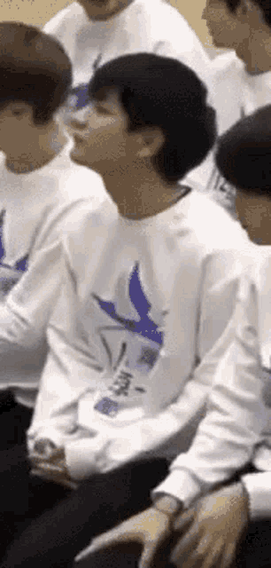 a man wearing a white shirt with a blue bird on it is sitting in a room with other people .