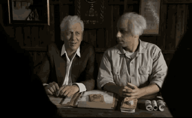 two men are sitting at a table with papers on it and one of them has a sign on the wall that says ' a '