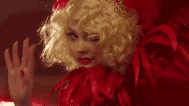 a woman in a red feathered dress and wig is waving .
