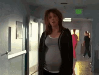 a pregnant woman walking down a hallway with a green exit sign on the wall