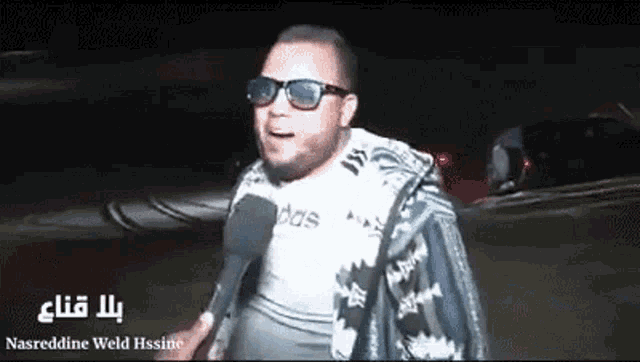 a man wearing sunglasses is holding a microphone and talking into it .