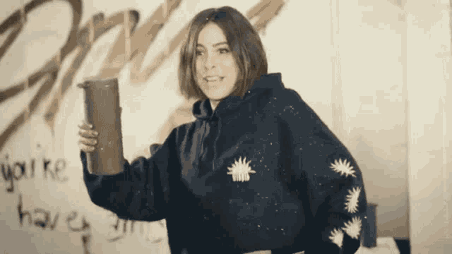 a woman in a black hoodie is holding a cup in front of a wall that says " i 'm going "