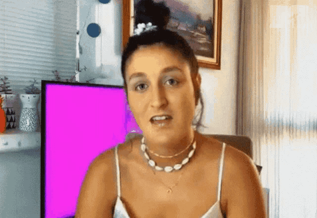 a woman wearing a necklace and a white tank top stands in front of a pink screen