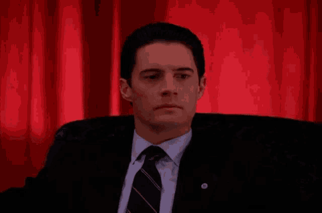 a man in a suit and tie is sitting in front of a red curtain and looking at the camera .