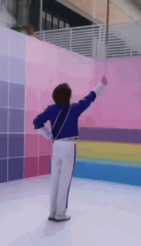 a person in a blue jacket and white pants is standing in front of a pink wall with a rainbow of colors .