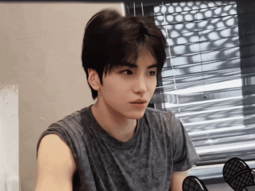 a young man in a grey shirt looks at the camera in front of a window