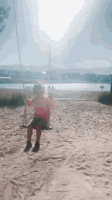 a little girl is sitting on a swing in the sand in front of a lake .
