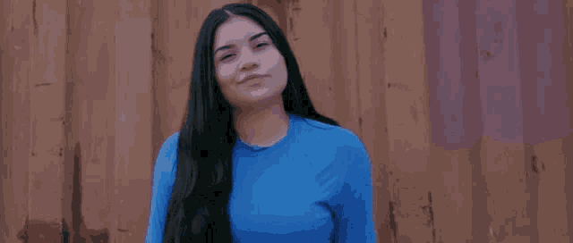 a woman with long black hair is wearing a blue shirt and standing in front of a wooden wall .