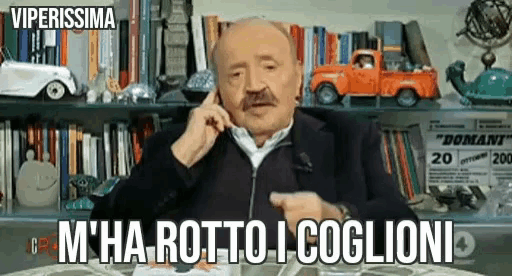 a man with a mustache is talking on a cell phone while sitting at a table in front of bookshelves .