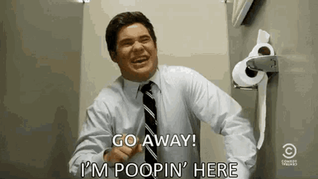 a man in a suit and tie is standing in a bathroom with a roll of toilet paper .