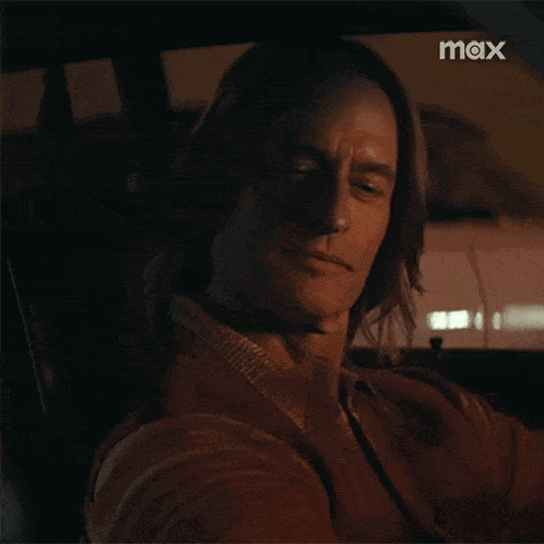 a man with long hair is sitting in the driver 's seat of a car with the word max on the bottom right