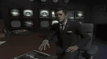 a man in a suit and tie sits at a desk in front of a wall of televisions with the letters cb on them