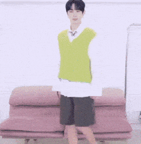 a man in a green vest and shorts is standing in front of a pink couch and giving a thumbs up .