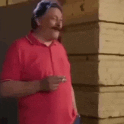a man in a red shirt is smoking a cigarette in front of a wooden building .