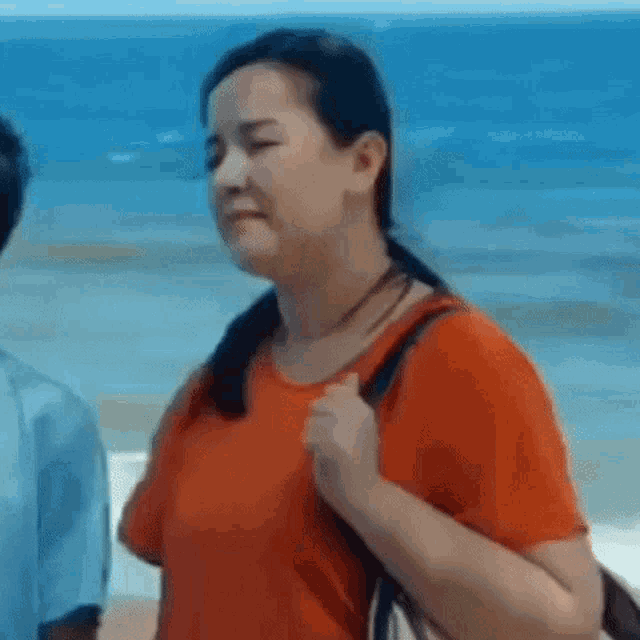 a woman in an orange shirt is standing on the beach holding a bag .