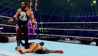 a wrestler is standing next to another wrestler who is laying on the ground in a wrestling ring .