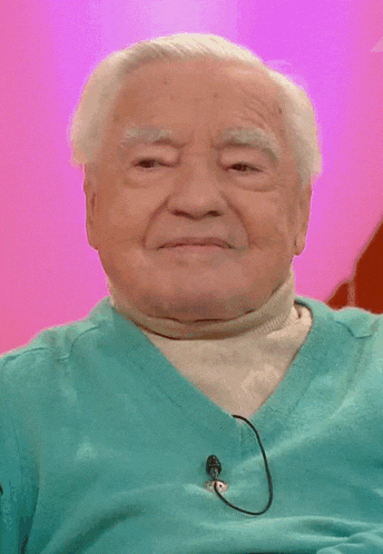 an elderly man wearing a blue sweater and a white turtleneck has a microphone attached to his neck