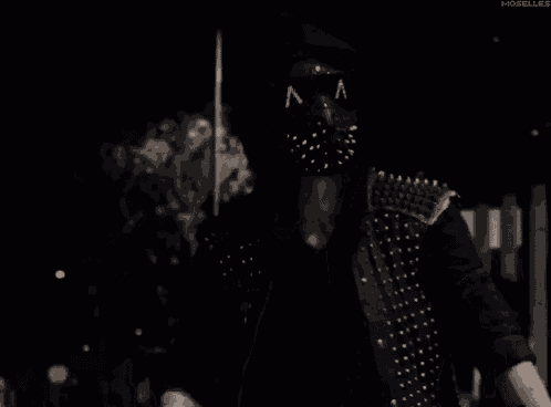 a man wearing a mask and a studded jacket is pointing at the camera .
