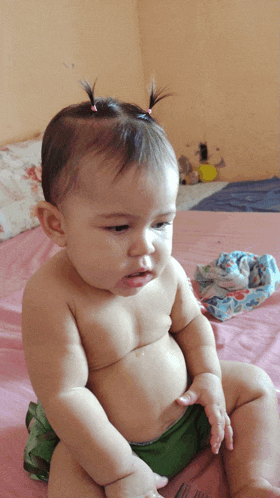 a baby without a shirt is sitting on a bed wearing green shorts