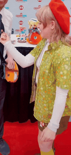 a woman in a green shirt and red beret is standing on a red carpet talking to a man