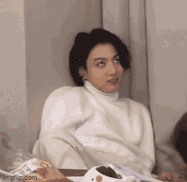 a young man in a white sweater is sitting on a bed with a turtleneck .