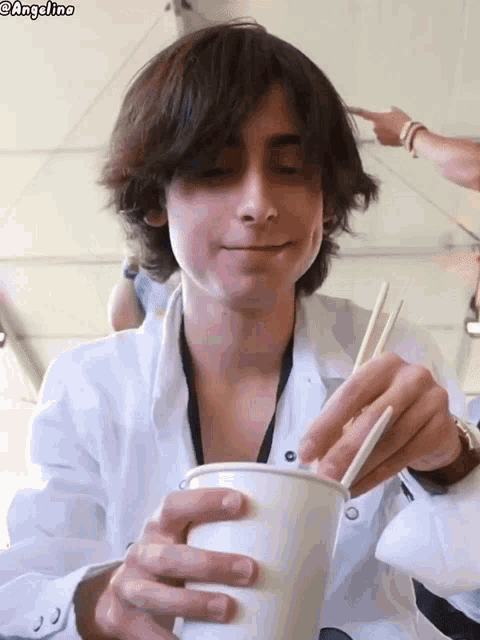a young man in a white shirt is holding a cup and chopsticks .